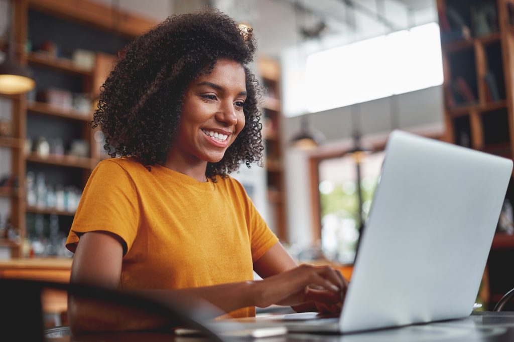 A woman with curly hair wearing a yellow shirt is smiling while working on a laptop in a café.