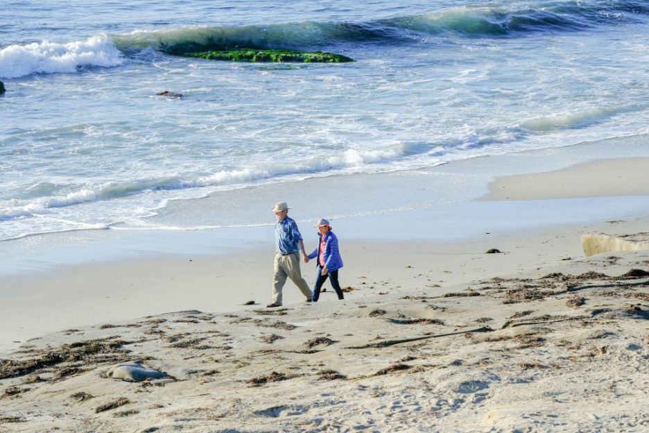An older couple holding hands and walking along a sandy beach with waves gently crashing in the background on a sunny day.