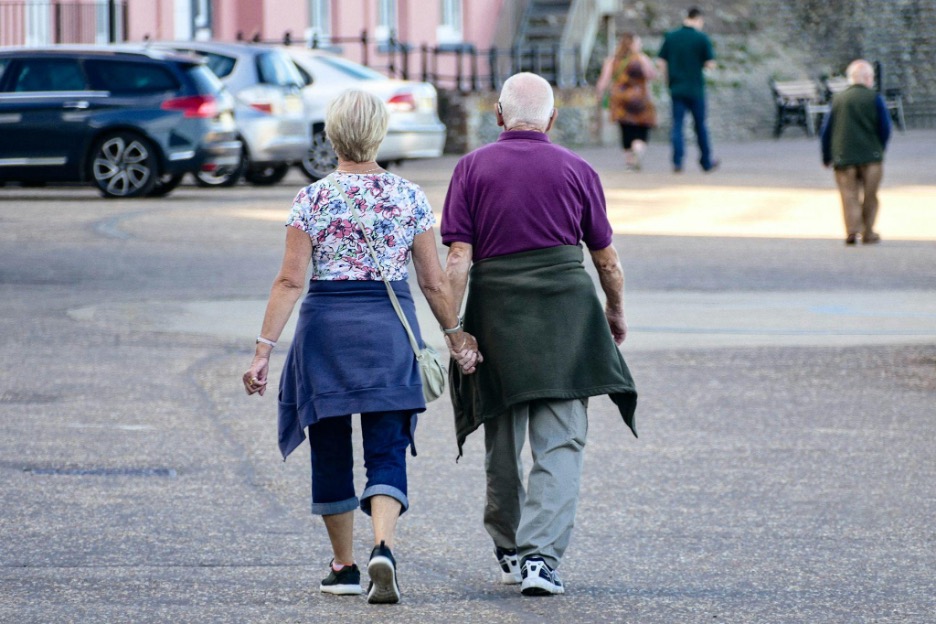 An elderly couple walks hand in hand down a paved street, casually dressed with sweaters tied around their waists, as other people stroll in the background.