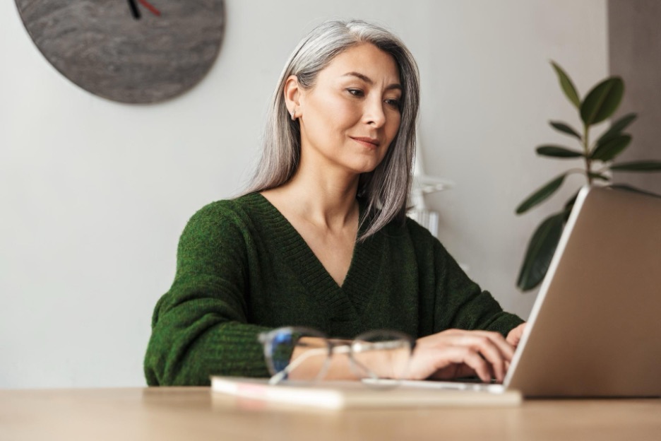 Older woman sits at a table looking at a laptop.