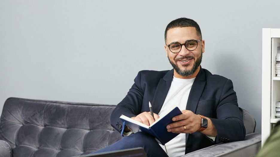 Smiling man in a navy blazer and glasses sitting on a gray couch, holding a notebook and pen.