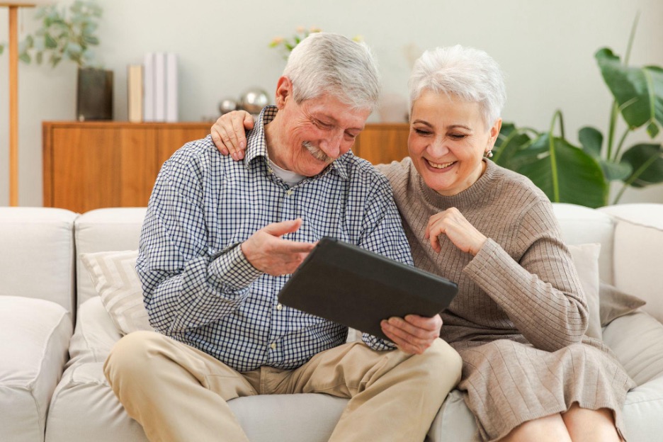 Smiling senior couple sitting on a couch looking at a tablet together, with the man pointing at the screen and the woman resting her hand on his shoulder.