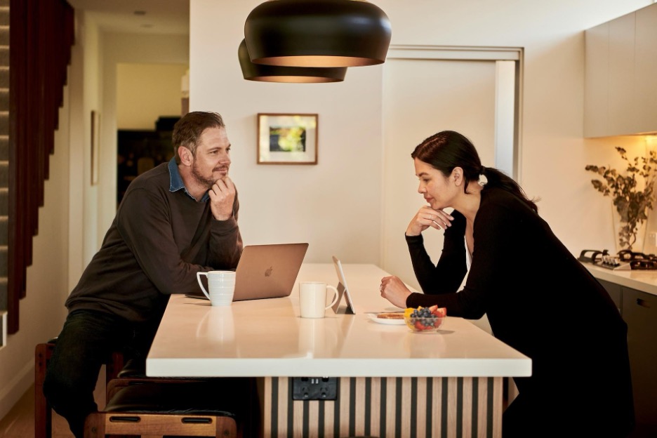 A man sits and a woman stands across from each other at a kitchen island, both looking at laptops.