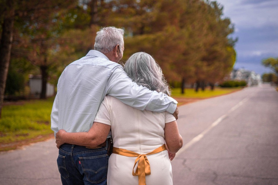 Older couple facing away with their arms around each other, walking down an open road.