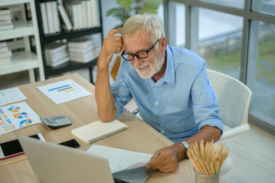An older man sits at a desk reviewing a document while working on his laptop, surrounded by charts, a calculator, and office supplies.