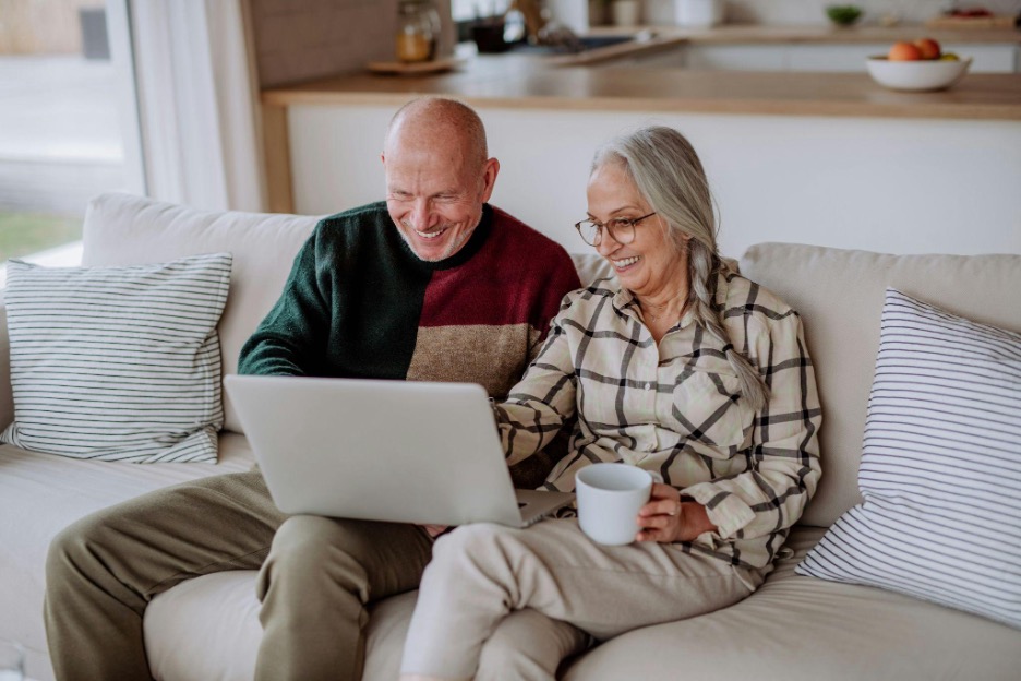 Elderly couple sitting on a couch with a laptop in their laps, pointing at the screen and smiling.
