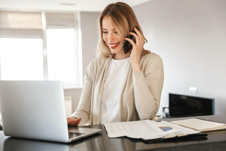 A smiling woman talks on the phone while working on her laptop, with documents and a notebook spread out in front of her.