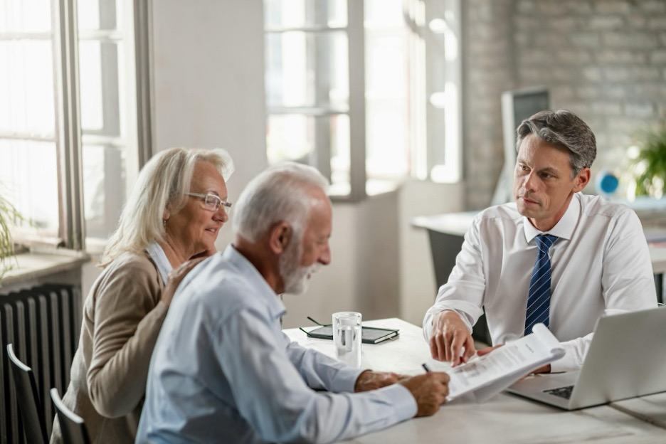 An elderly couple meets with a person in a bright office, reviewing and signing documents together.