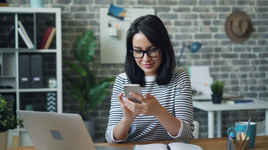 Woman sitting at a desk looking at a smartphone in her hands.