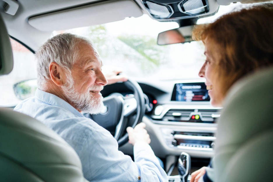 An elderly couple smiles at each other in the front seats of a car.