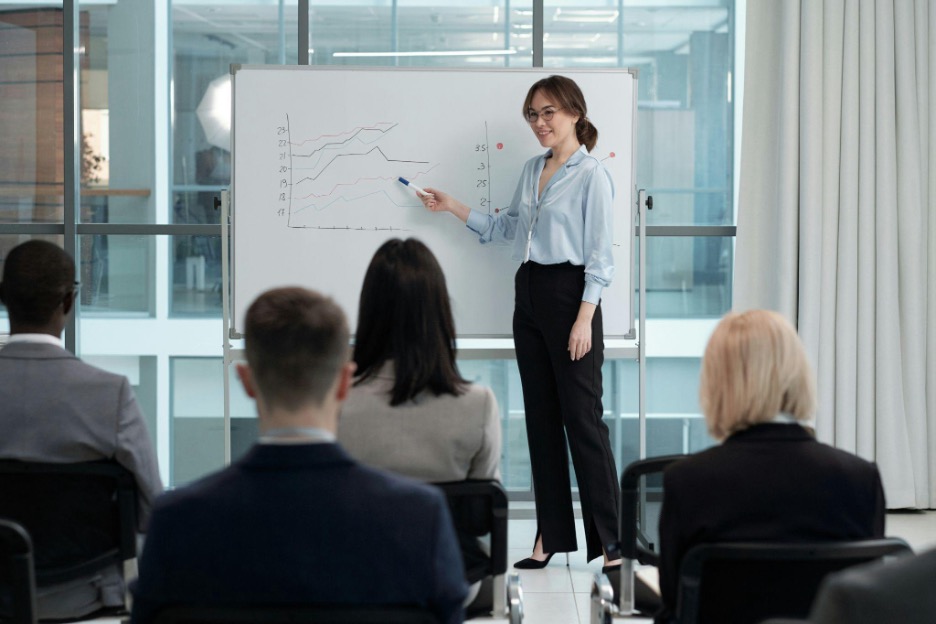 A woman speaks in front of a group, pointing at a line graph on a whiteboard.